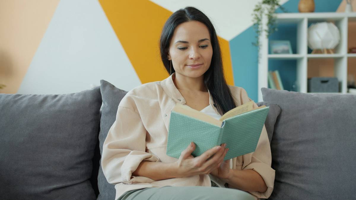 Good-looking young woman is reading book and smiling then turning page sitting on comfortable sofa alone in nice apartment. Youth, literature and lifestyle concept.
